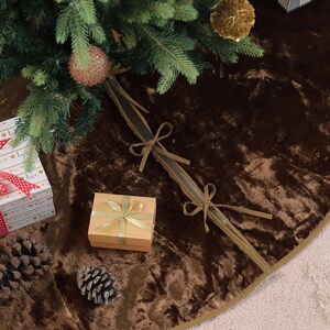 Puede incluir: Una falda de &aacute;rbol de Navidad de terciopelo marr&oacute;n con ribete dorado, decorada con una caja de regalo, pi&ntilde;as y un regalo envuelto. La falda del &aacute;rbol est&aacute; debajo de un &aacute;rbol de Navidad con adornos.