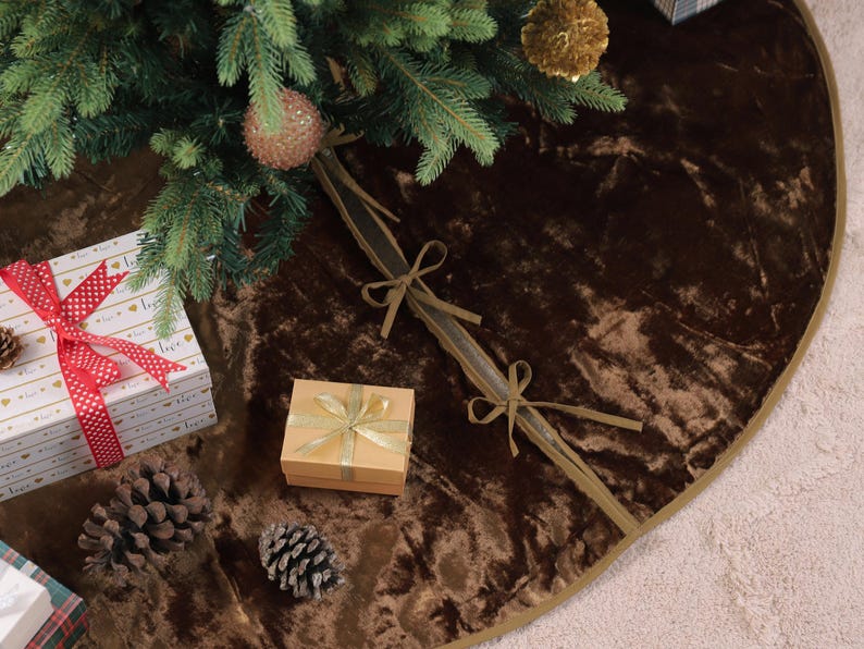 Puede incluir: Una falda de &aacute;rbol de Navidad de terciopelo marr&oacute;n con ribete dorado, decorada con una caja de regalo, pi&ntilde;as y un regalo envuelto. La falda del &aacute;rbol est&aacute; debajo de un &aacute;rbol de Navidad con adornos.