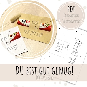 Könnte beinhalten: Eine druckbare PDF-Vorlage mit dem deutschen Text "du bist gut genug!", einem kleinen Herzsymbol und einem Punktemuster. Das Design ist um einen Schokoriegel gewickelt dargestellt, ergänzt durch weiße Papierbögen mit demselben Text.