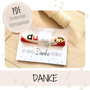 以下が含まれることがあります： 「Danke」の文字とチョコレートバーが描かれた白いカード。カードには「ein dickes Danke verdient!」の文字と小さなハートが描かれています。背景には紐のロールがあります。画像には「PDF Druckvorlage Sofortdownload」の文字も含まれています。