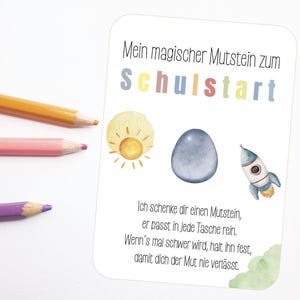 May include: A white card with the text "Mein magischer Mutstein zum Schulstart" and a watercolor illustration of a sun, a blue egg, and a rocket. The card also has text in German. Three colored pencils are in the image.