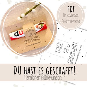 以下が含まれることがあります： 「du hast es geschafft!」と「Herzlichen Glückwunsch!」の文字が入ったチョコレートバー付きクラフト紙のギフトカード。カードには小さなタグと、白い先端の緑の小枝が飾られています。