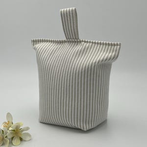 May include: A grey and white striped fabric doorstop with a matching handle. The doorstop is a rectangular shape and is made of a soft, textured fabric. The doorstop is designed to hold a door open.