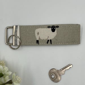 May include: A key fob featuring a sheep design. The key fob is made of light grey fabric, with a black and white sheep illustration. A silver metal key ring and a key are also visible.