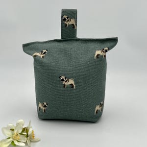 May include: A green fabric doorstop with embroidered pug dog patterns. The doorstop has a handle at the top and is decorated with multiple pug dog illustrations. A decorative and functional home accessory.