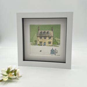 May include: A framed watercolour print depicting a yellow house with a green roof and a blue door. The artwork is set against a green backdrop and framed in a white square frame. A small sign reads "Bee Hotel".