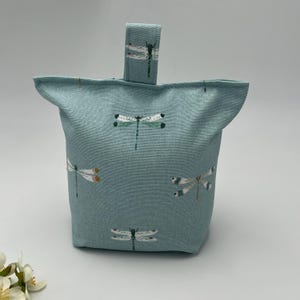 May include: A teal fabric doorstop with a dragonfly pattern. The doorstop is rectangular with a small handle. The dragonfly design includes white, black, and orange accents. The doorstop is designed to hold a door open, a practical home accessory.