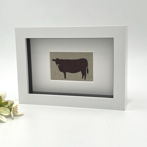 May include: A framed picture with a brown cow silhouette on a beige background. The cow has a pink detail on its face. The artwork is set in a white frame with a black interior border, suitable for farmhouse or country decor.