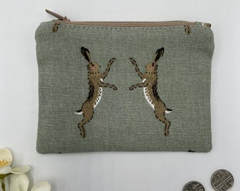 May include: A small, rectangular, sage green zippered pouch featuring two brown hares in a dancing pose. The hares have white bellies and are set against a textured fabric background. The zipper is a light brown color.