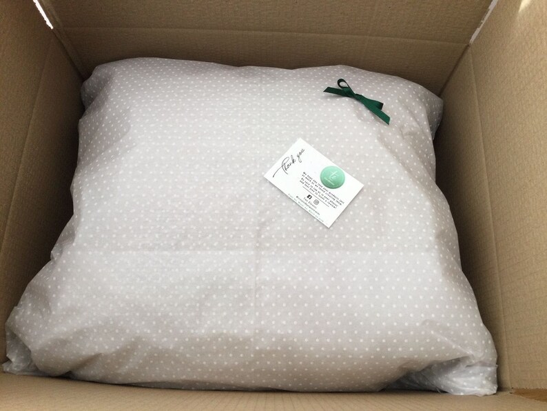 May include: A white tissue paper with small white dots, folded into a square shape, inside a brown cardboard box. A green ribbon is tied around the tissue paper. A small white card with the text "Thank you" and a logo is placed on top of the tissue paper.