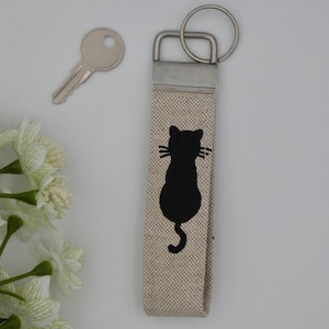 May include: A beige fabric keyring with a black silhouette of a cat sitting on its hind legs. The keyring has a silver metal ring and a silver metal clasp.