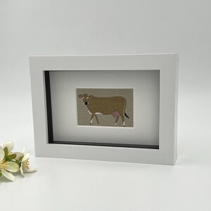 May include: A framed picture of a brown cow with a pink udder and white legs, set against a textured tan background. The artwork is enclosed in a white rectangular frame with a black inner border. A charming piece of farm animal decor, suitable for a child's room.