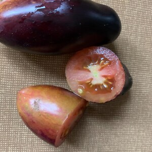 Op de afbeelding: Drie erfgoedtomaten, één is diep paars, één is lichtpaars met een roze tint en één is doormidden gesneden en laat een rood interieur zien.