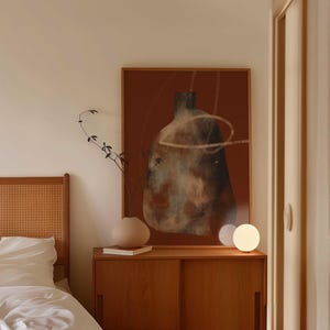 May include: A bedside table with a wooden top and two drawers. A white ceramic lamp sits on the table next to a brown vase with a leafy branch. A framed abstract painting with brown and white colors hangs on the wall behind the table.