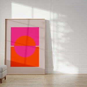 May include: A framed abstract art print with a pink and orange geometric design. The print features a large circle divided in half by a thin white line. The top half of the circle is pink, and the bottom half is orange. The circle is set against a white background.