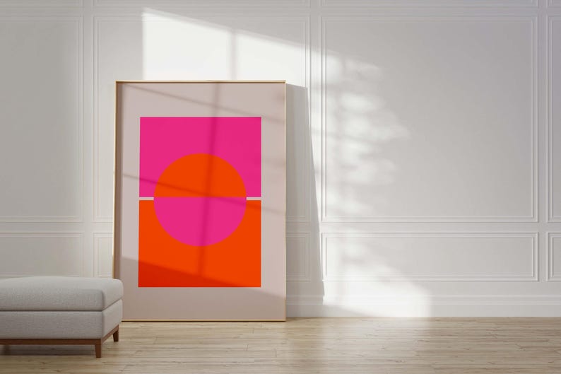 May include: A framed abstract art print with a pink and orange geometric design. The print features a large circle divided in half by a thin white line. The top half of the circle is pink, and the bottom half is orange. The circle is set against a white background.