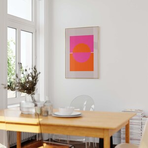 May include: A framed abstract art print with a pink and orange geometric design. The print is hanging on a white wall in a dining room with a wooden table and clear chairs.