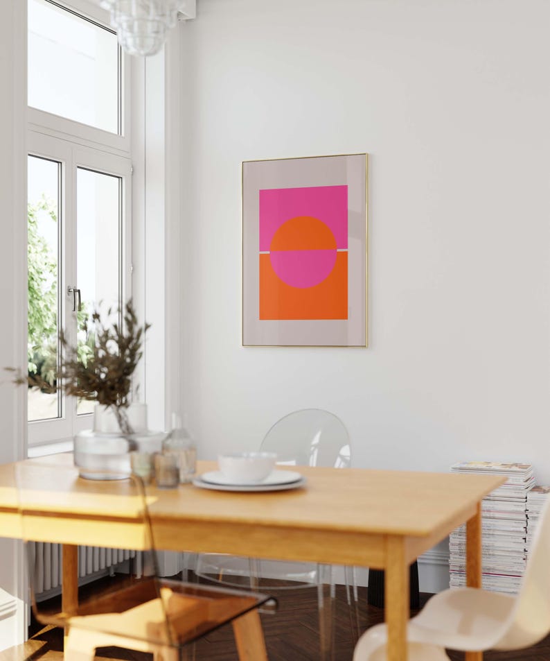 May include: A framed abstract art print with a pink and orange geometric design. The print is hanging on a white wall in a dining room with a wooden table and clear chairs.
