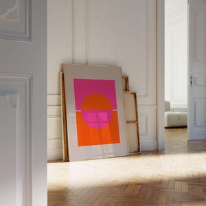 May include: A framed abstract art print with a pink and orange geometric design. The print is in a gold frame and is leaning against a white wall.