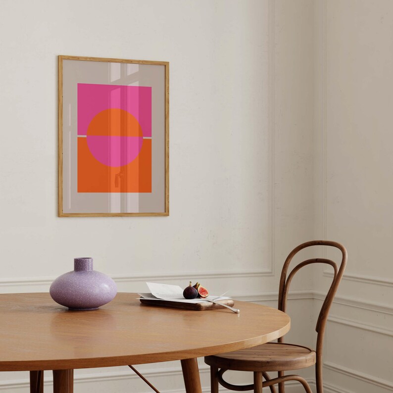 May include: A framed abstract print with a pink and orange geometric design. The print is hanging on a white wall above a wooden table with a purple vase and a wooden chair.