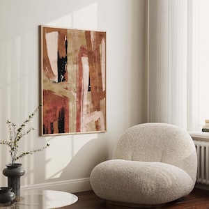 Puede incluir: Impresión de arte abstracto en tonos cálidos de marrón, rosa y beige con acentos negros. La impresión está enmarcada y colgada en una pared blanca sobre un sillón blanco.