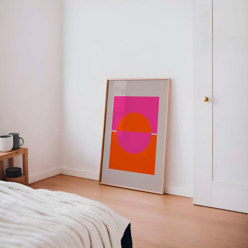 May include: A framed print with a geometric design featuring a pink and orange circle divided by a thin white line. The print is in a light wood frame and is hanging on a white wall.