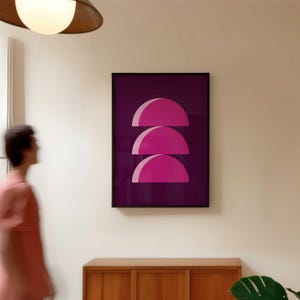 May include: A framed art print featuring three stacked, semi-circular shapes in shades of pink against a deep purple background. The artwork is displayed on a white wall, above a wooden cabinet.