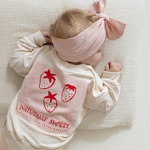 Op de afbeelding: Een baby in een crèmekleurig shirt met lange mouwen met een roze vierkante patch met drie rode aardbeien en de tekst "NATURALLY SWEET". De baby draagt ook een bijpassende roze hoofdband. De baby ligt op een crèmekleurig kussen.