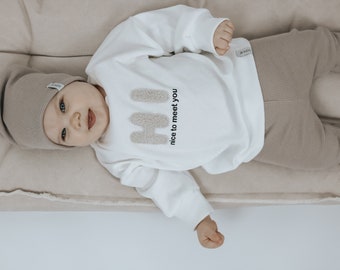 May include: A baby wearing a white sweater with the text "Hi nice to meet you" and brown pants. The baby is wearing a brown knit beanie.