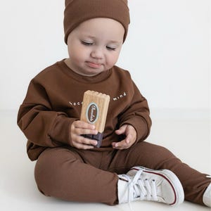 Puede incluir: Un beb&eacute; con gorro, sudadera y pantalones de punto acanalado marr&oacute;n. El beb&eacute; sostiene un juguete de madera con la etiqueta "Chocolate". El conjunto se combina con zapatillas blancas con detalles rojos. La sudadera tiene las palabras "chocolate mind" impresas.