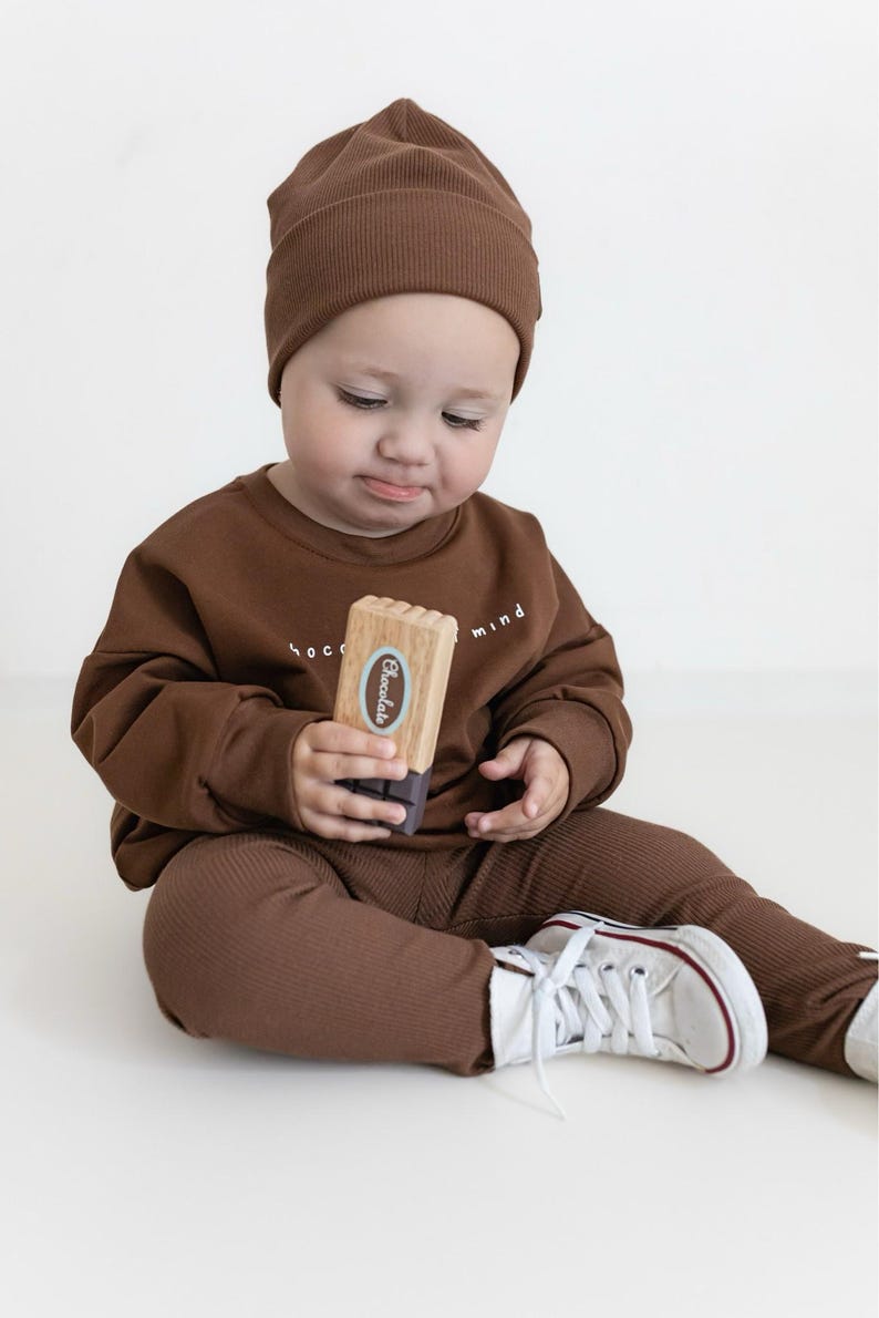 Puede incluir: Un beb&eacute; con gorro, sudadera y pantalones de punto acanalado marr&oacute;n. El beb&eacute; sostiene un juguete de madera con la etiqueta "Chocolate". El conjunto se combina con zapatillas blancas con detalles rojos. La sudadera tiene las palabras "chocolate mind" impresas.