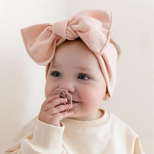 Puede incluir: Un beb&eacute; con una diadema de lazo rosa claro y un chupete a juego. La diadema est&aacute; hecha de una tela suave y texturizada. El beb&eacute; lleva una camisa de manga larga de color crema.