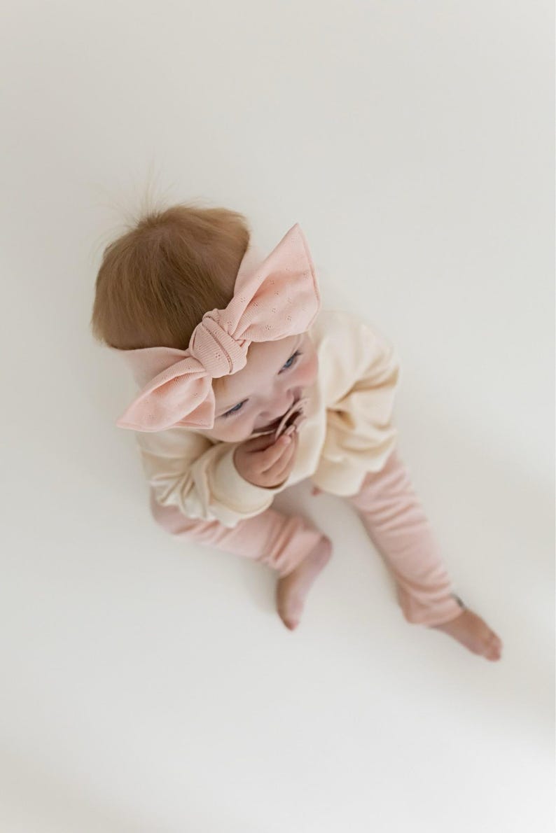 Puede incluir: Un beb&eacute; con una diadema de lazo rosa claro, una camisa de manga larga color crema y pantalones rosas claros. El beb&eacute; est&aacute; acostado sobre una superficie blanca.