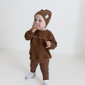Puede incluir: Un ni&ntilde;o peque&ntilde;o con un conjunto marr&oacute;n de gorro, sudadera y leggings, con el texto "chace state of mind". El ni&ntilde;o tambi&eacute;n lleva zapatillas blancas con ribete rojo. El conjunto es a juego.