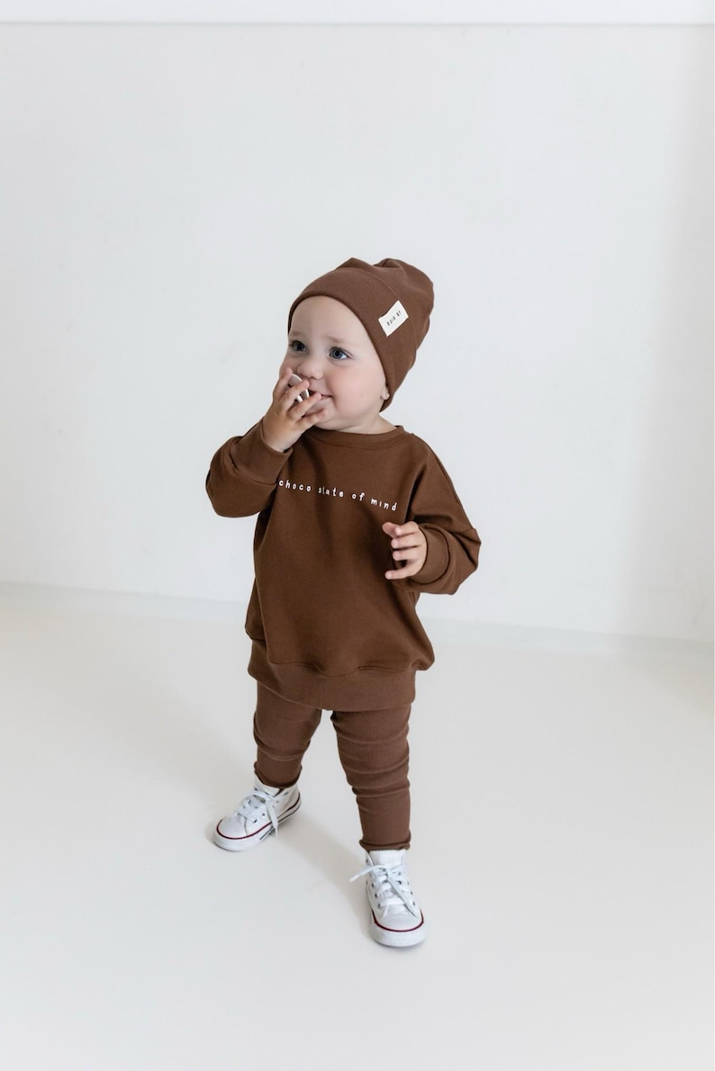 Puede incluir: Un ni&ntilde;o peque&ntilde;o con un conjunto marr&oacute;n de gorro, sudadera y leggings, con el texto "chace state of mind". El ni&ntilde;o tambi&eacute;n lleva zapatillas blancas con ribete rojo. El conjunto es a juego.