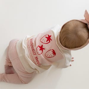 May include: A baby wearing a light pink outfit, including pants and a long-sleeved shirt with a strawberry design and the text "NATURALLY SWEET". A matching pink bow headband completes the ensemble.