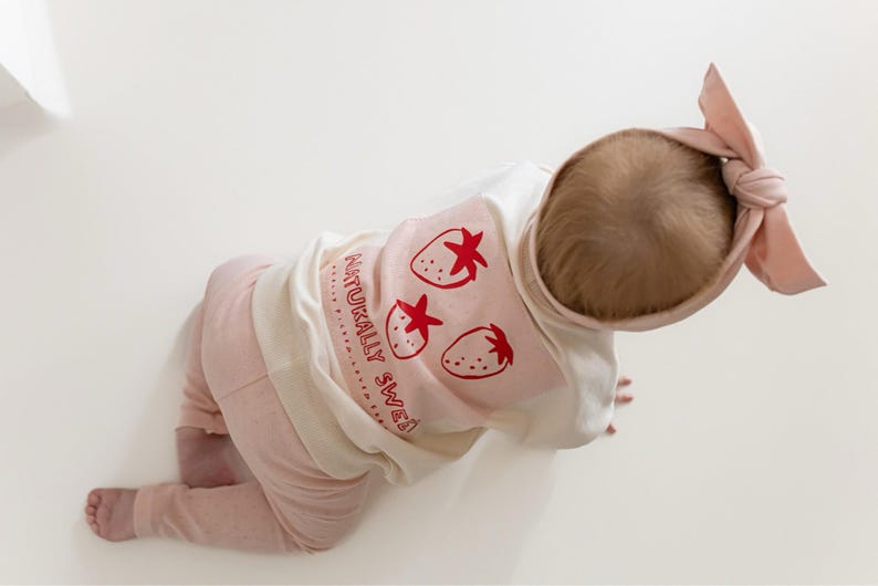 May include: A baby wearing a light pink outfit, including pants and a long-sleeved shirt with a strawberry design and the text "NATURALLY SWEET". A matching pink bow headband completes the ensemble.