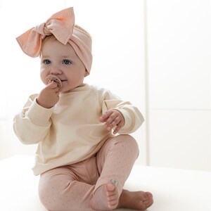 Puede incluir: Un beb&eacute; con una sudadera crema y leggings rosas con un sutil estampado de lunares. El beb&eacute; tambi&eacute;n lleva una diadema rosa a juego con un lazo grande. El beb&eacute; sostiene un chupete y est&aacute; sentado sobre una superficie blanca.