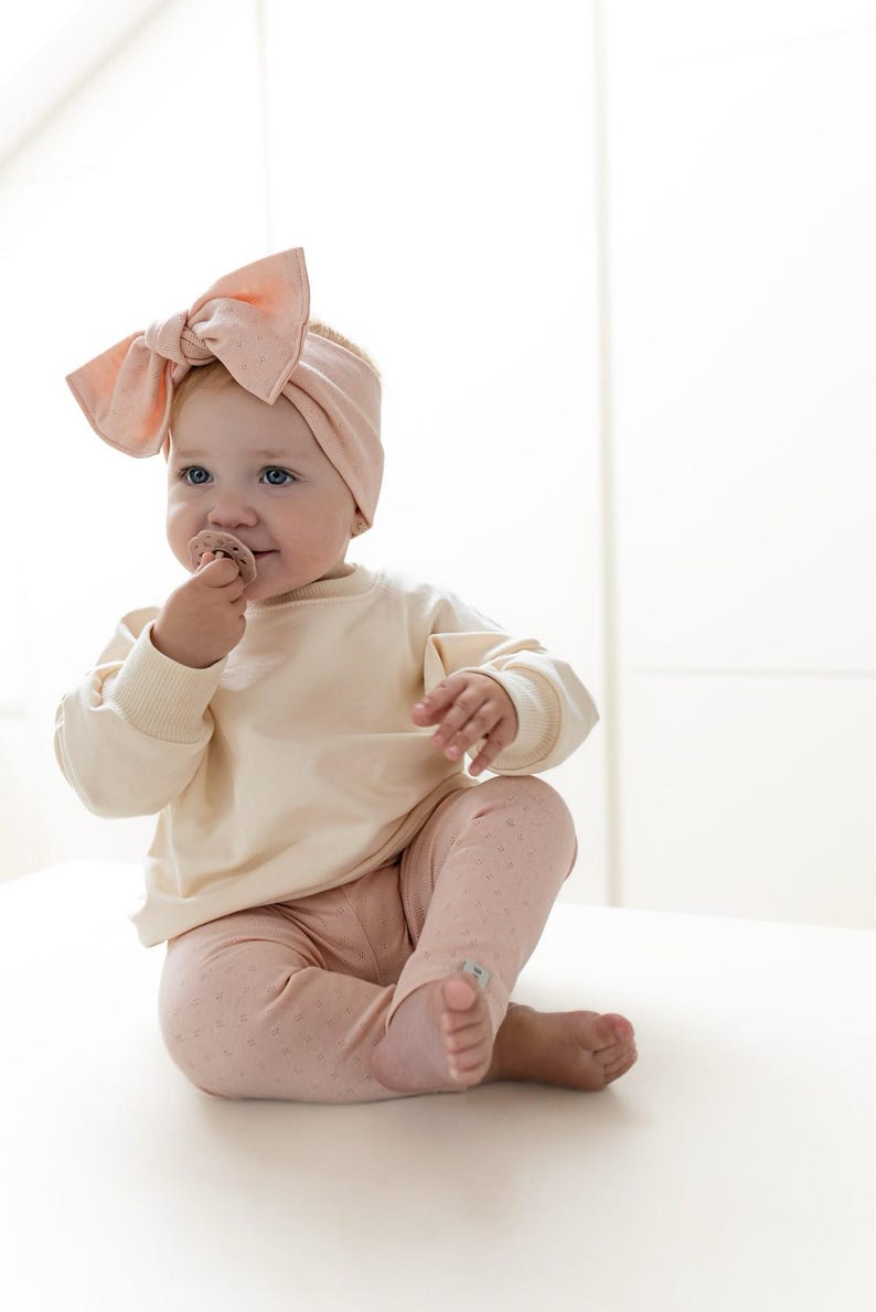 Puede incluir: Un beb&eacute; con una sudadera crema y leggings rosas con un sutil estampado de lunares. El beb&eacute; tambi&eacute;n lleva una diadema rosa a juego con un lazo grande. El beb&eacute; sostiene un chupete y est&aacute; sentado sobre una superficie blanca.