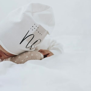 Könnte beinhalten: Eine weiße Baby-Mütze mit dem Aufdruck "The answer is sleep". Die Mütze sitzt auf dem Kopf eines Babys.