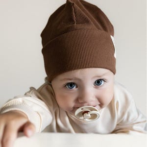 Könnte beinhalten: Ein Baby mit einer braunen Rippstrickmütze und einem Schnuller. Das Baby hat leuchtend blaue Augen und trägt ein cremefarbenes Langarmshirt. Auf der Seite der Mütze befindet sich ein kleines weißes Etikett.