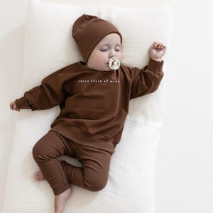 Puede incluir: Un beb&eacute; dormido con un conjunto marr&oacute;n que incluye gorro, sudadera y pantal&oacute;n. La sudadera tiene el texto blanco "choco state of mind". El beb&eacute; est&aacute; acostado sobre una almohada blanca y tiene un chupete en la boca.