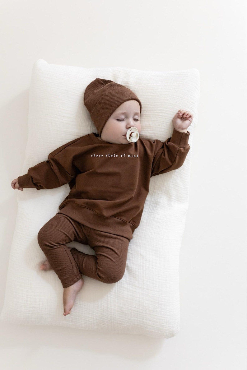Puede incluir: Un beb&eacute; dormido con un conjunto marr&oacute;n que incluye gorro, sudadera y pantal&oacute;n. La sudadera tiene el texto blanco "choco state of mind". El beb&eacute; est&aacute; acostado sobre una almohada blanca y tiene un chupete en la boca.