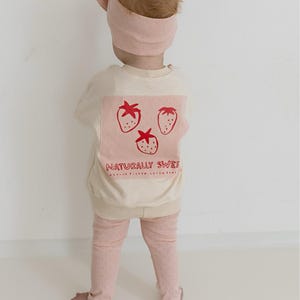 May include: A child wearing a light pink headband and matching outfit. The cream-colored sweatshirt features a pink square with three red strawberry illustrations and the text "NATURALLY SWEET". The leggings are light pink with small pink dots.