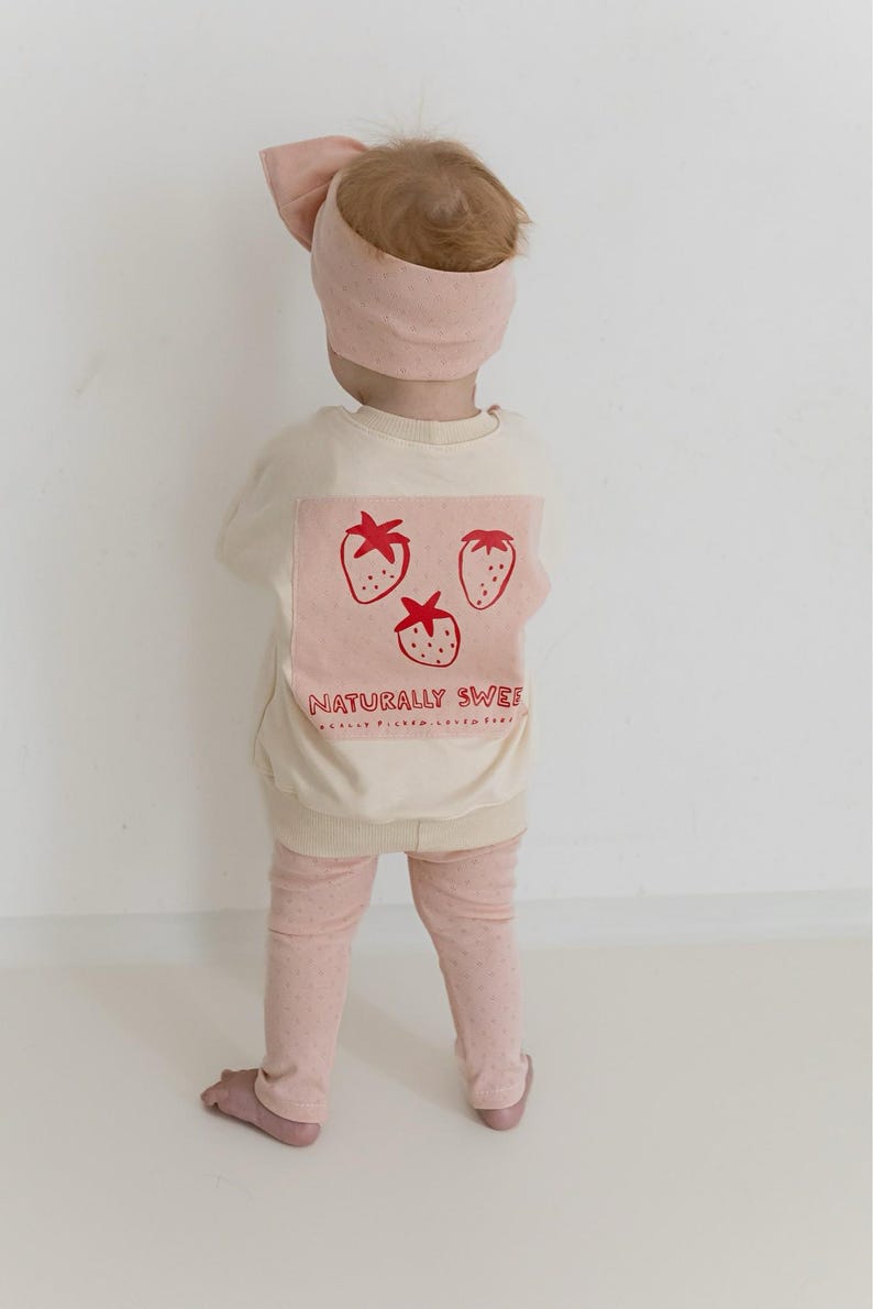 May include: A child wearing a light pink headband and matching outfit. The cream-colored sweatshirt features a pink square with three red strawberry illustrations and the text "NATURALLY SWEET". The leggings are light pink with small pink dots.