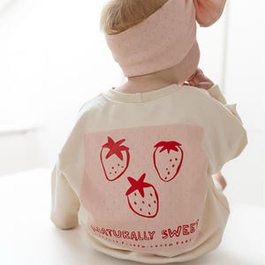 May include: A baby wearing a cream-colored sweatshirt and a matching pink headband. The sweatshirt features a pink square with three red strawberry illustrations and the text "NATURALLY SWEET".