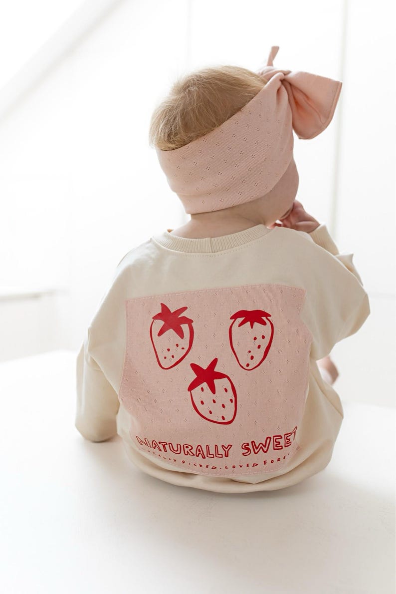 May include: A baby wearing a cream-colored sweatshirt and a matching pink headband. The sweatshirt features a pink square with three red strawberry illustrations and the text "NATURALLY SWEET".