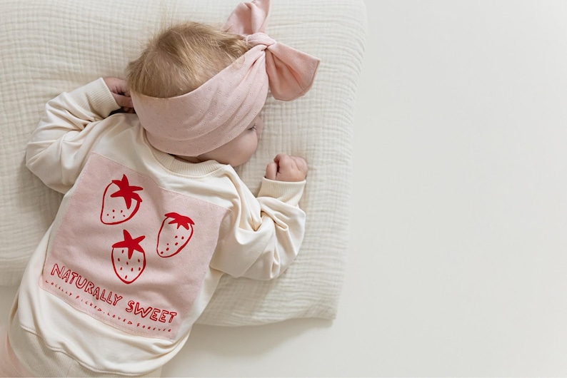 May include: A baby wearing a cream-colored long-sleeved shirt with a pink square patch featuring three red strawberries and the text "NATURALLY SWEET." The baby also wears a matching pink headband. The baby is lying on a cream-colored pillow.
