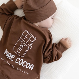 Puede incluir: Conjunto de ropa de beb&eacute; marr&oacute;n, que incluye una camiseta de manga larga y un gorro. La camiseta presenta un gr&aacute;fico blanco de una tableta de chocolate y las palabras "PURE COCOA melts hearts - not snow". El conjunto est&aacute; dise&ntilde;ado para la comodidad y el estilo.