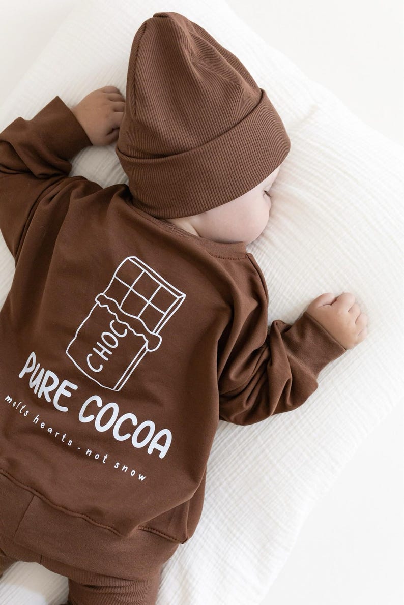 Puede incluir: Conjunto de ropa de beb&eacute; marr&oacute;n, que incluye una camiseta de manga larga y un gorro. La camiseta presenta un gr&aacute;fico blanco de una tableta de chocolate y las palabras "PURE COCOA melts hearts - not snow". El conjunto est&aacute; dise&ntilde;ado para la comodidad y el estilo.